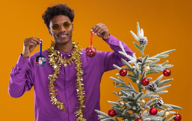 Sorridente giovane afro-americano con gli occhiali con la ghirlanda di orpelli intorno al collo in piedi vicino all'albero di Natale decorato su sfondo arancione