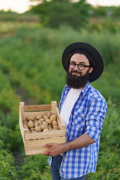 Sorridente giovane agricoltore che tiene una cassa di legno di patate sul campo di patate verdi