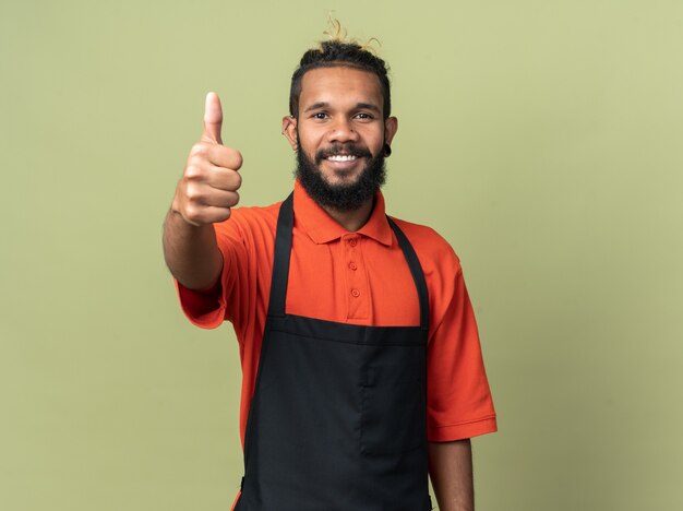 Sorridente giovane barbiere afroamericano che indossa l'uniforme che mostra il pollice in su