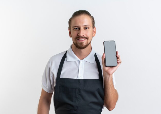 Sorridente giovane barbiere bello che indossa uniforme che mostra telefono cellulare isolato sul muro bianco