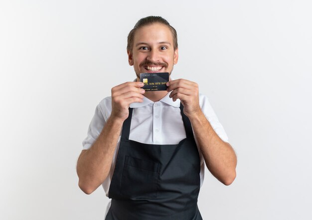 Sorridente giovane barbiere bello indossando l'uniforme che mostra la carta di credito isolata sul muro bianco