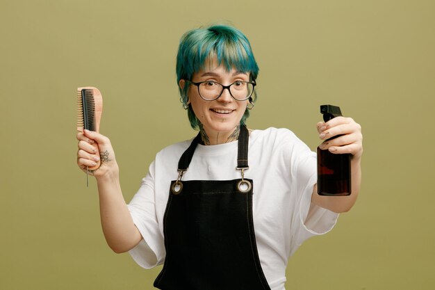 Sorridente giovane barbiere femminile che indossa occhiali uniforme che tiene pettini guardando la fotocamera allungando la lacca per capelli verso la fotocamera isolata su sfondo verde oliva