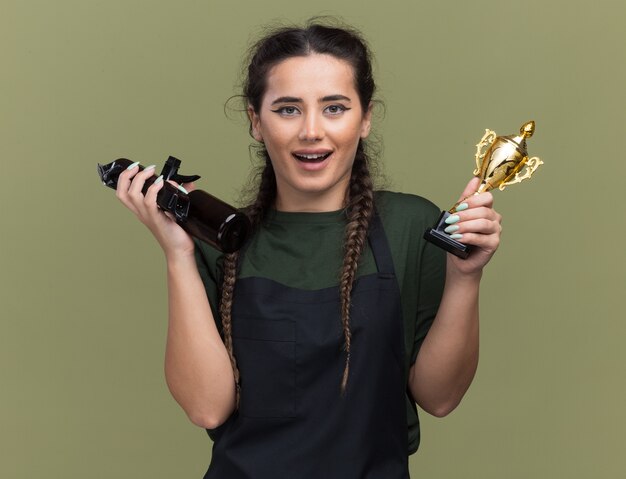 Sorridente giovane barbiere femminile in uniforme che tiene tazza del vincitore con tagliacapelli isolato su parete verde oliva