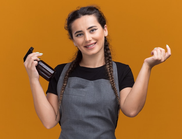 Sorridente giovane barbiere femminile in uniforme con flacone spray che sparge la mano isolata sulla parete arancione