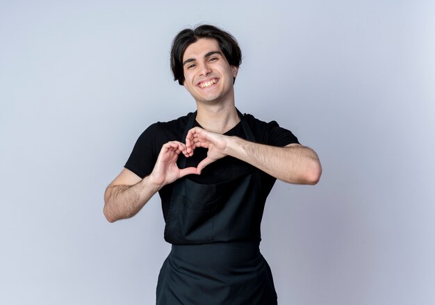 Sorridente giovane barbiere maschio bello in uniforme che mostra il gesto del cuore isolato su bianco