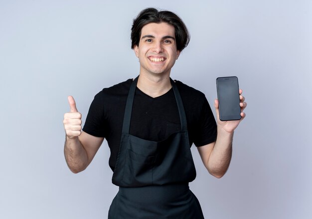 Sorridente giovane barbiere maschio bello in uniforme che tiene il telefono con il pollice in alto isolato sulla parete bianca