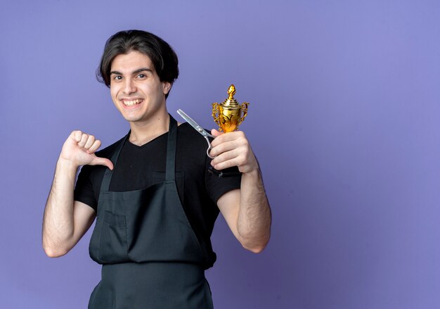 Sorridente giovane barbiere maschio bello in uniforme che tiene la tazza del vincitore e indica se stesso isolato sull'azzurro con lo spazio della copia