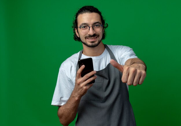 Sorridente giovane barbiere maschio caucasico indossando l'uniforme e occhiali tenendo il telefono cellulare e mostrando il pollice in alto isolato su sfondo verde con spazio di copia