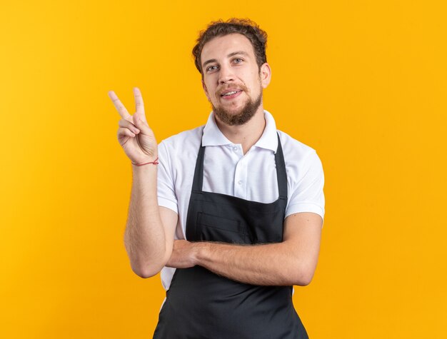 Sorridente giovane barbiere maschio che indossa l'uniforme che mostra gesto di pace isolato su sfondo giallo