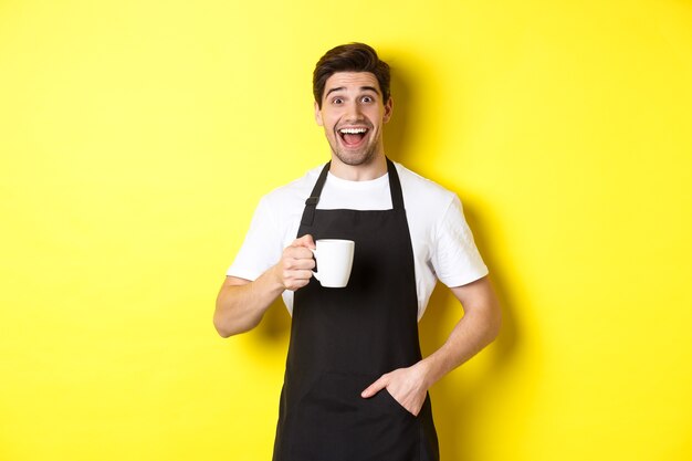 Sorridente giovane barista in grembiule nero tenendo la tazza di caffè, in piedi su sfondo giallo.