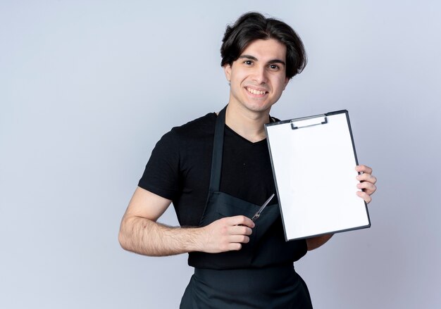 Sorridente giovane bel barbiere maschio in uniforme che tiene appunti con le forbici isolate su bianco