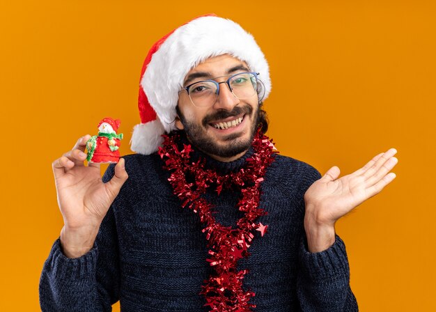 Sorridente giovane bel ragazzo che indossa il cappello di Natale con la ghirlanda sul collo tenendo il giocattolo diffondendo la mano isolata su sfondo arancione