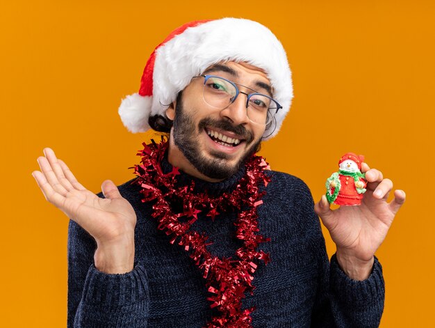 Sorridente giovane bel ragazzo che indossa il cappello di Natale con la ghirlanda sul collo tenendo il giocattolo diffondendo la mano isolata su sfondo arancione