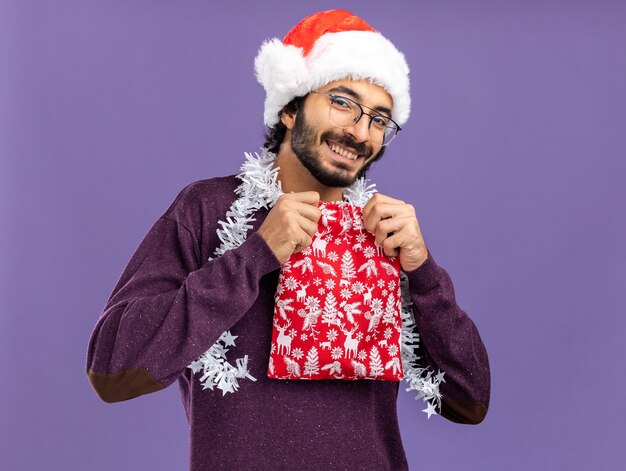 Sorridente giovane bel ragazzo che indossa il cappello di Natale con la ghirlanda sul collo tenendo il sacchetto di Natale isolato su sfondo blu
