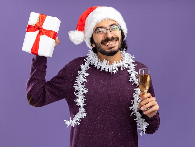 Sorridente giovane bel ragazzo che indossa il cappello di Natale con la ghirlanda sul collo tenendo la confezione regalo con un bicchiere di champagne isolato su sfondo blu