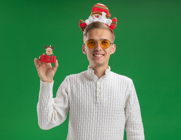 Foto gratuito sorridente giovane bel ragazzo che indossa la fascia di babbo natale con gli occhiali che tengono il giocattolo di renne con la data che sembra isolata sul muro verde