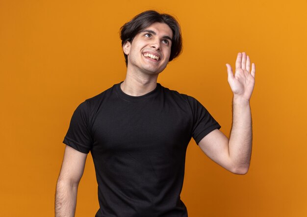 Sorridente giovane bel ragazzo che indossa la maglietta nera che mostra ciao gesto isolato sulla parete arancione