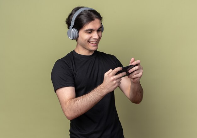 Sorridente giovane bel ragazzo che indossa la maglietta nera e cuffie che giocano gioco sul telefono isolato sulla parete verde oliva