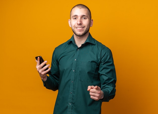 Sorridente giovane bel ragazzo che indossa una camicia verde tenendo il telefono e indicando davanti