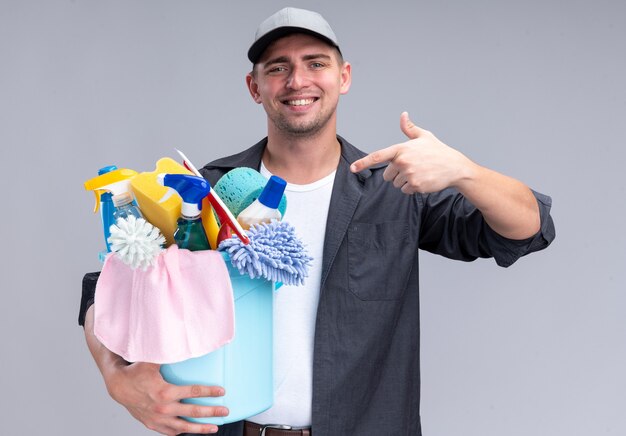 Sorridente giovane bel ragazzo delle pulizie che indossa t-shirt e berretto che tiene e punti al secchio di strumenti di pulizia isolato sulla parete bianca