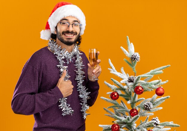 Sorridente giovane bel ragazzo in piedi vicino albero di Natale che indossa il cappello di Natale con la ghirlanda sul collo tenendo un bicchiere di champagne che mostra il pollice in alto isolato su sfondo arancione