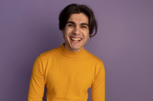 Sorridente giovane bel ragazzo indossa maglione dolcevita giallo isolato sulla parete viola con spazio di copia