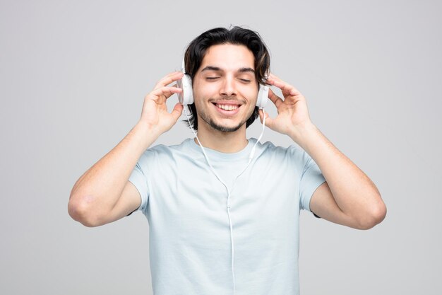 sorridente giovane bell'uomo che indossa le cuffie afferrandole godendosi la musica con gli occhi chiusi isolati su sfondo bianco