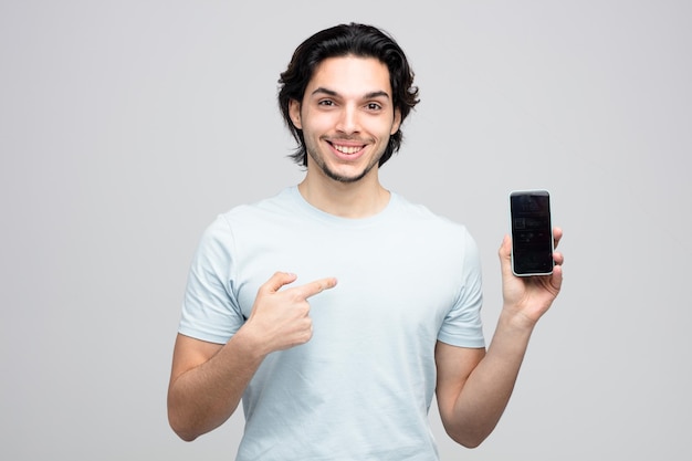 sorridente giovane bell'uomo che mostra il telefono cellulare che punta ad esso guardando la fotocamera isolata su sfondo bianco