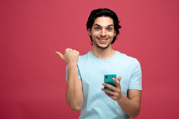 sorridente giovane bell'uomo che tiene il telefono cellulare guardando la fotocamera che punta al lato isolato su sfondo rosso con spazio di copia