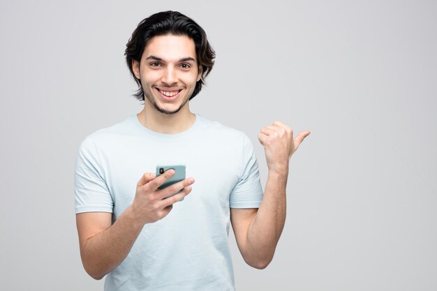 sorridente giovane bell'uomo in possesso di telefono cellulare guardando la fotocamera che punta al lato isolato su sfondo bianco con spazio di copia