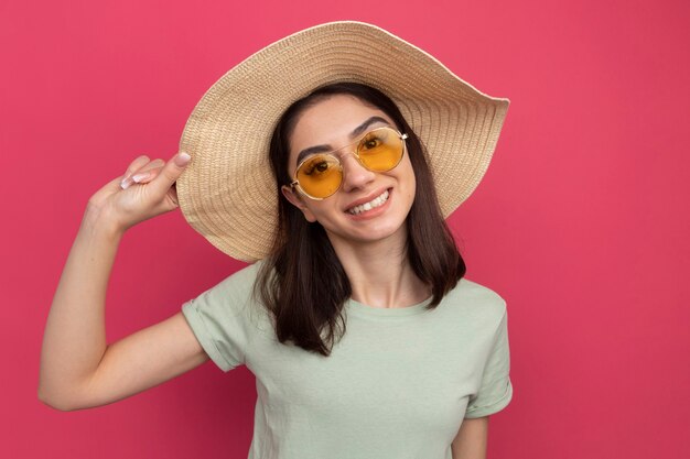 Sorridente giovane bella donna caucasica che indossa cappello da spiaggia e occhiali da sole che afferra il cappello