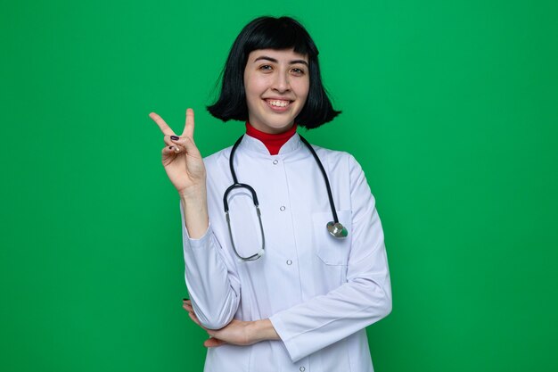 Sorridente giovane bella donna caucasica in uniforme da medico con lo stetoscopio che gesturing segno di vittoria