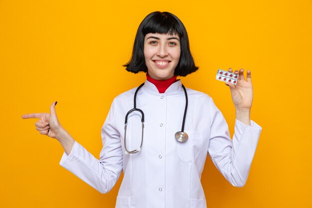 Sorridente giovane bella donna caucasica in uniforme da medico con stetoscopio che tiene la confezione della pillola e che punta a lato