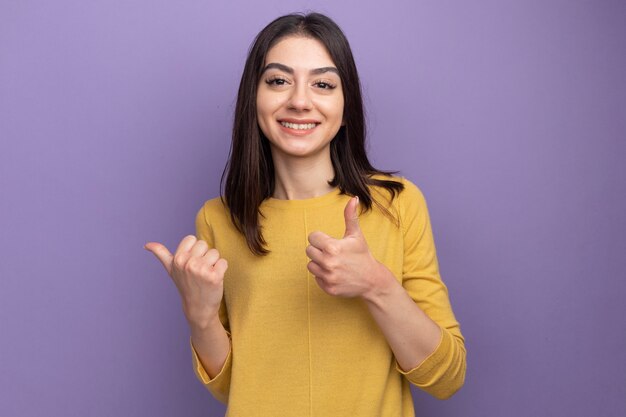 Sorridente giovane bella donna che guarda la parte anteriore che punta a lato mostrando pollice in alto isolato sul muro viola con spazio copia