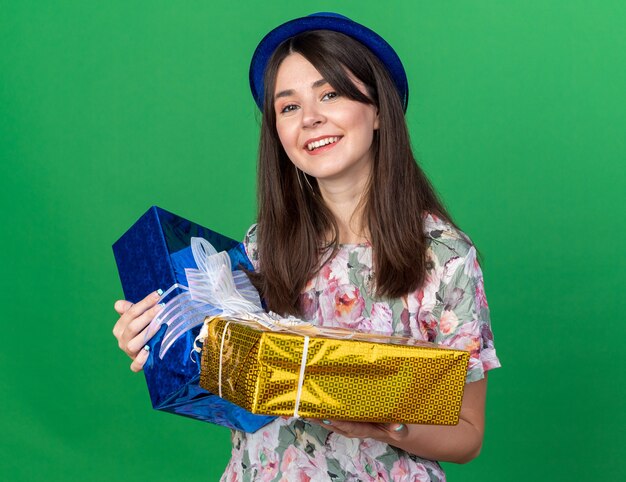 Sorridente giovane bella donna che indossa cappello da festa tenendo scatole regalo isolate su parete verde