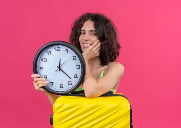 Sorridente giovane bella donna viaggiatore che tiene la valigia e l'orologio sulla parete rosa isolata
