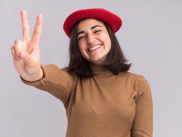 Sorridente giovane bella ragazza caucasica con berretto che gesticola segno di vittoria victory