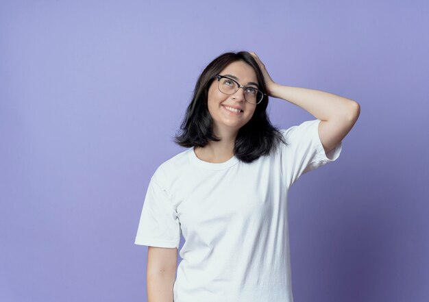 Sorridente giovane bella ragazza caucasica con gli occhiali cercando con la mano sulla testa isolata su sfondo viola con spazio di copia