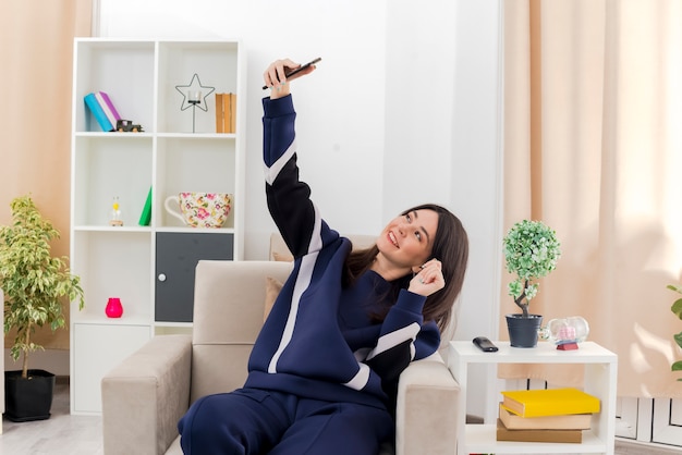 Sorridente giovane bella ragazza caucasica seduto sulla poltrona nel soggiorno progettato prendendo selfie e tenendo la mano in aria