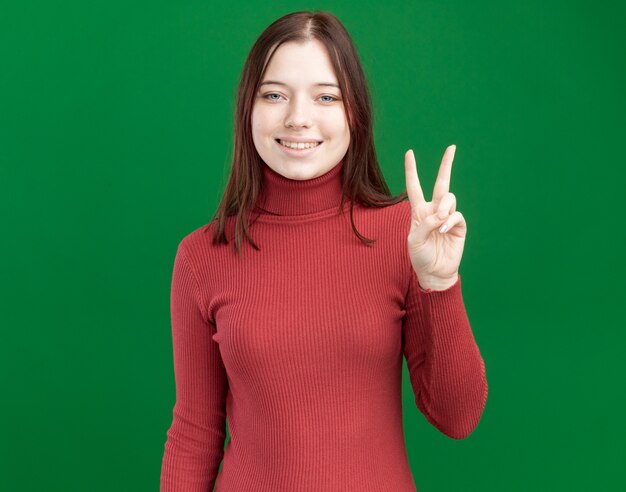 Sorridente giovane bella ragazza che fa segno di pace isolato sulla parete verde con spazio di copia
