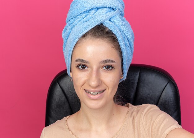 Sorridente giovane bella ragazza che indossa bretelle dentali capelli avvolti in asciugamano tenendo la fotocamera isolata su sfondo rosa