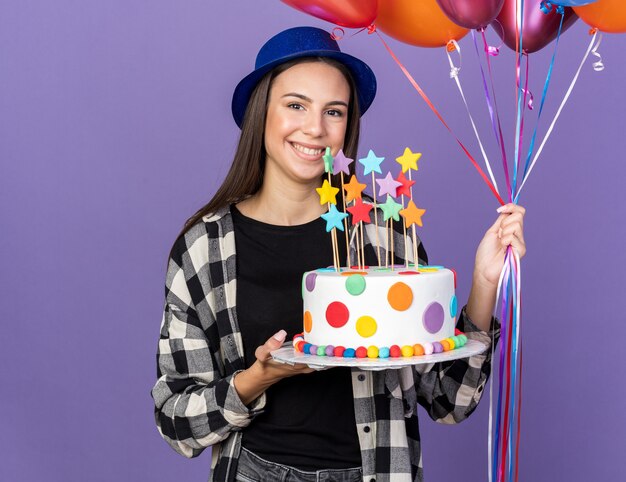 Sorridente giovane bella ragazza che indossa cappello da festa tenendo palloncini con torta isolata sulla parete blu