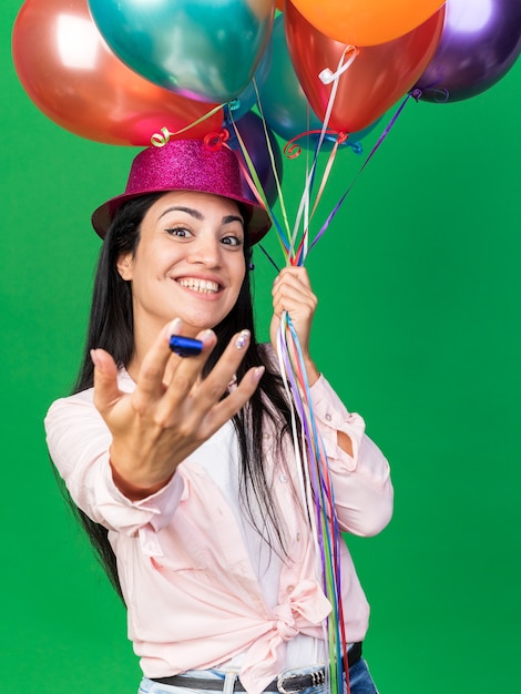 Sorridente giovane bella ragazza che indossa cappello da festa tenendo palloncini e porgendo fischietto da festa isolato su parete verde