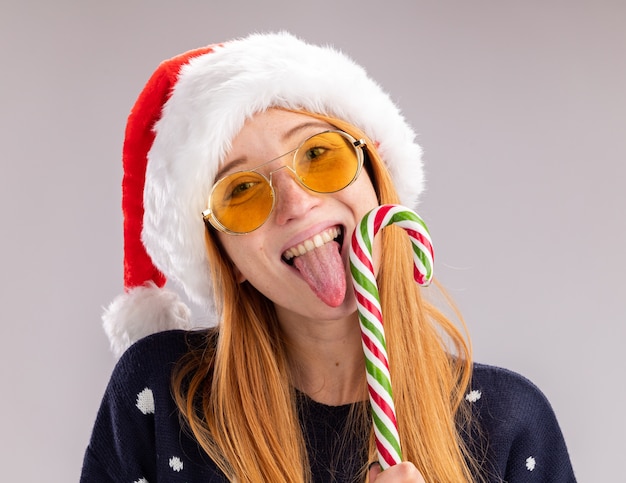 Sorridente giovane bella ragazza che indossa cappello e occhiali di natale che tengono e leccano caramelle di natale isolate sul muro bianco