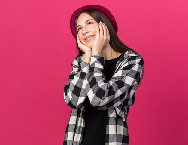 Sorridente giovane bella ragazza che indossa il cappello da festa coperto le guance sul viso isolato sul muro rosa