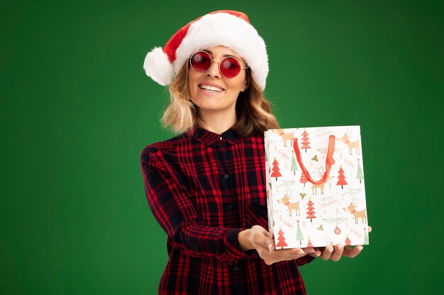 Sorridente giovane bella ragazza che indossa il cappello di Natale con gli occhiali che tengono fuori la borsa regalo in telecamera isolata su sfondo verde