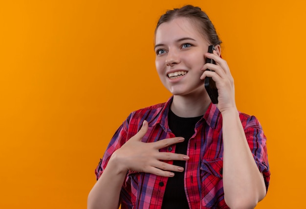 Sorridente giovane bella ragazza che indossa la camicia rossa parla al telefono su sfondo giallo isolato con spazio di copia