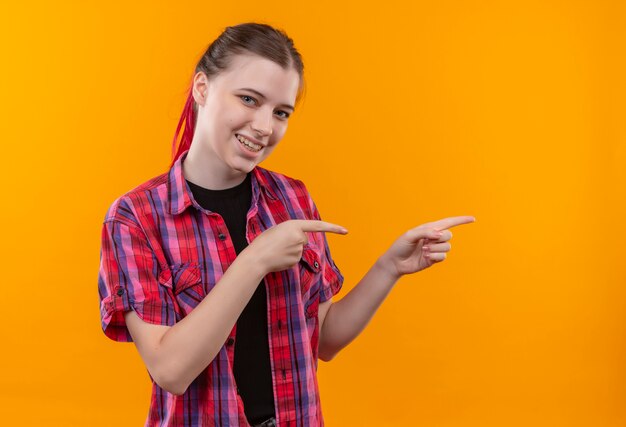 Sorridente giovane bella ragazza che indossa la camicia rossa punta le dita a lato su sfondo giallo isolato con spazio di copia