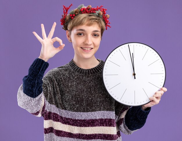 Sorridente giovane bella ragazza che indossa la corona di testa di Natale che tiene l'orologio che guarda l'obbiettivo che fa segno giusto isolato su fondo viola