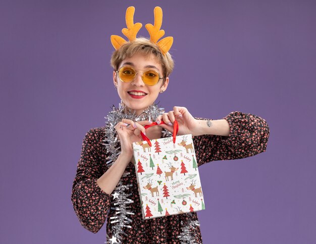 Sorridente giovane bella ragazza che indossa la fascia di corna di renna e la ghirlanda di orpelli intorno al collo con gli occhiali che tengono il sacchetto del regalo di Natale che guarda l'obbiettivo isolato su priorità bassa viola